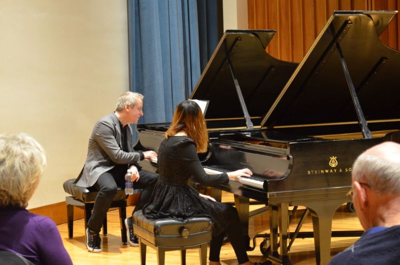A man and woman each sit at their own piano while he coaches the student musician.