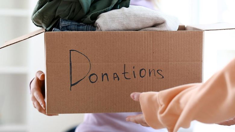 A person handing over a cardboard box containing clothes, with "Donations" written on it.
