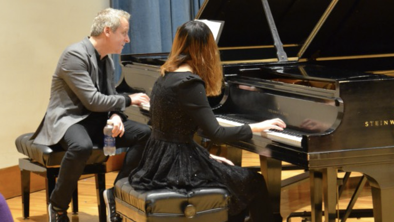A man and woman each sit at their own piano while he coaches the student musician.
