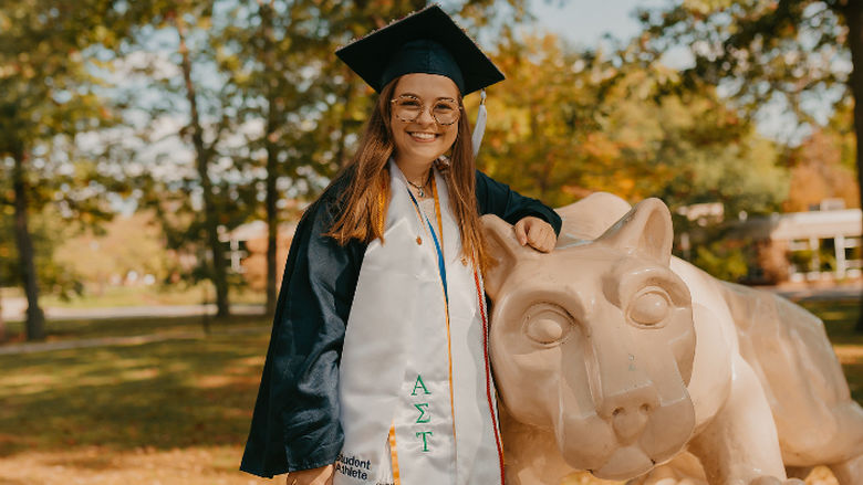 Woman poses in graduation cap and gown with a Lion Shrine statue
