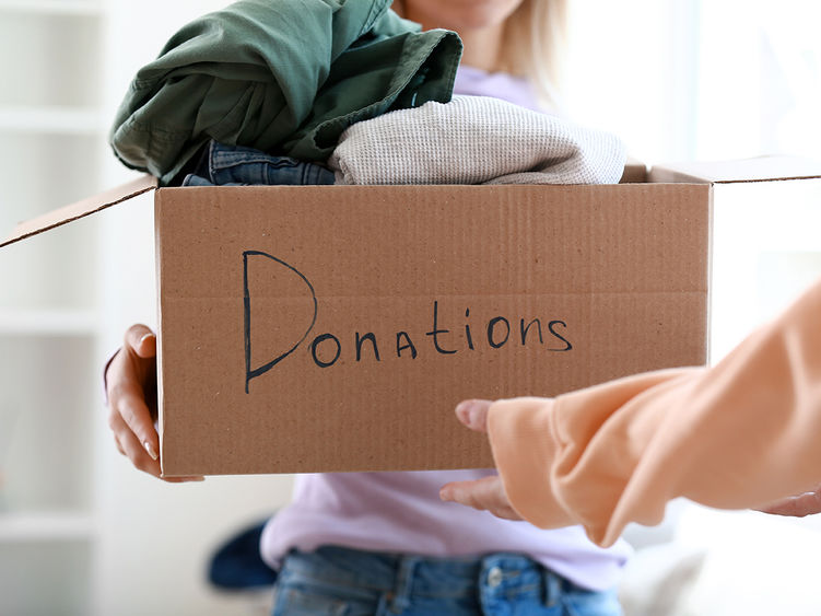 A person handing over a cardboard box containing clothes, with "Donations" written on it.