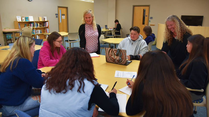 A group of students sits around a table in a library or classroom setting, engaged in discussion and writing. Two instructors stand with them, smiling and interacting. Bookshelves, chairs, and a clock are visible in the background, suggesting an academic or collaborative learning environment.
