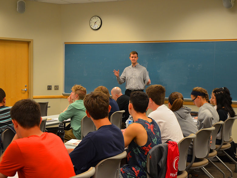 A male instructor stands at the front of a classroom, speaking to a group of college students seated at desks facing him.