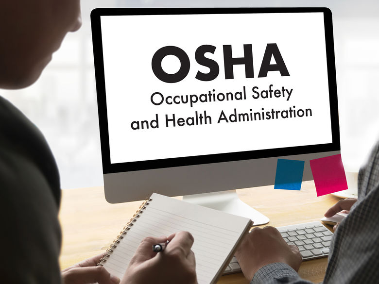 Two people sitting at a desk in front of a computer monitor displaying the text OSHA Occupational Safety and Health Administration. One person is writing in a notebook while the other uses a keyboard. The monitor has pink and blue sticky notes attached to it.