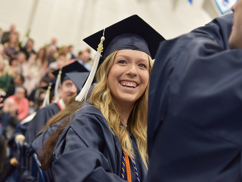 A smiling graduate wearing a navy blue cap and gown with a white tassel sits among a crowd during a commencement ceremony, surrounded by fellow graduates and an audience in the background.