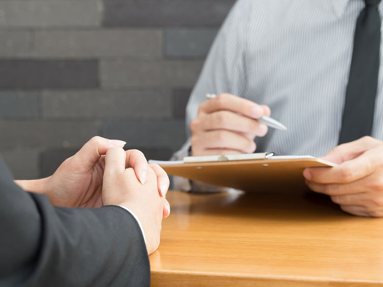 A close-up of two people in a professional setting, with one holding a clipboard and pen, while the other has hands clasped, likely during an interview or meeting.