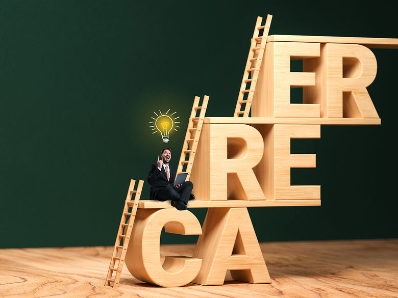 A man in a suit sits on large, wooden block letters spelling CAREER, holding a laptop, with ladders leaning against the letters and a lightbulb above his head.