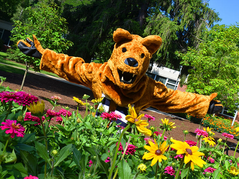 The Nittany Lion holding out his arms in a welcoming way behind a bed of flowers.