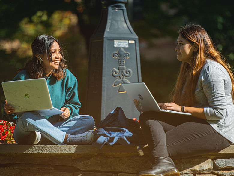 Two students sitting around the campus clock on their laptops
