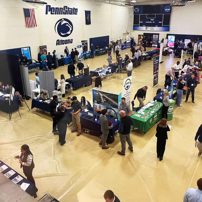Overhead view of a career fair inside a gymnasium at Penn State Altoona, with numerous employer booths set up and students and representatives engaging in conversations. The Penn State Altoona logo is prominently displayed on the back wall, along with American and Pennsylvania state flags.