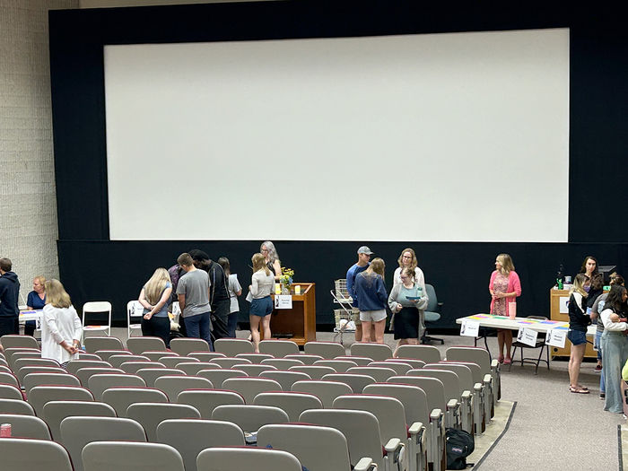 People gather at various tables in a large lecture hall, engaging with exhibitors beneath a large blank screen.