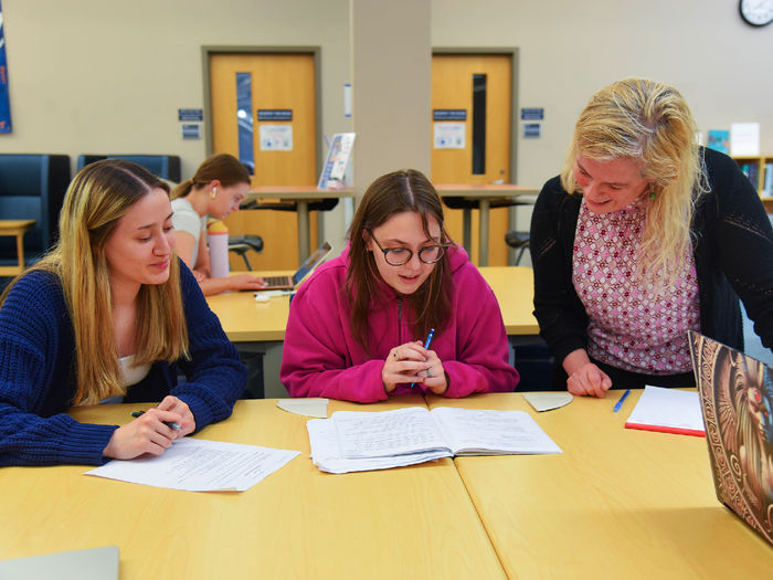 A student in a pink hoodie reads from an open workbook while seated between another student and an instructor. The instructor leans in to offer support, smiling. A third student listens attentively, holding a pen. The setting appears to be a library or study space with bookshelves and computers in the background.