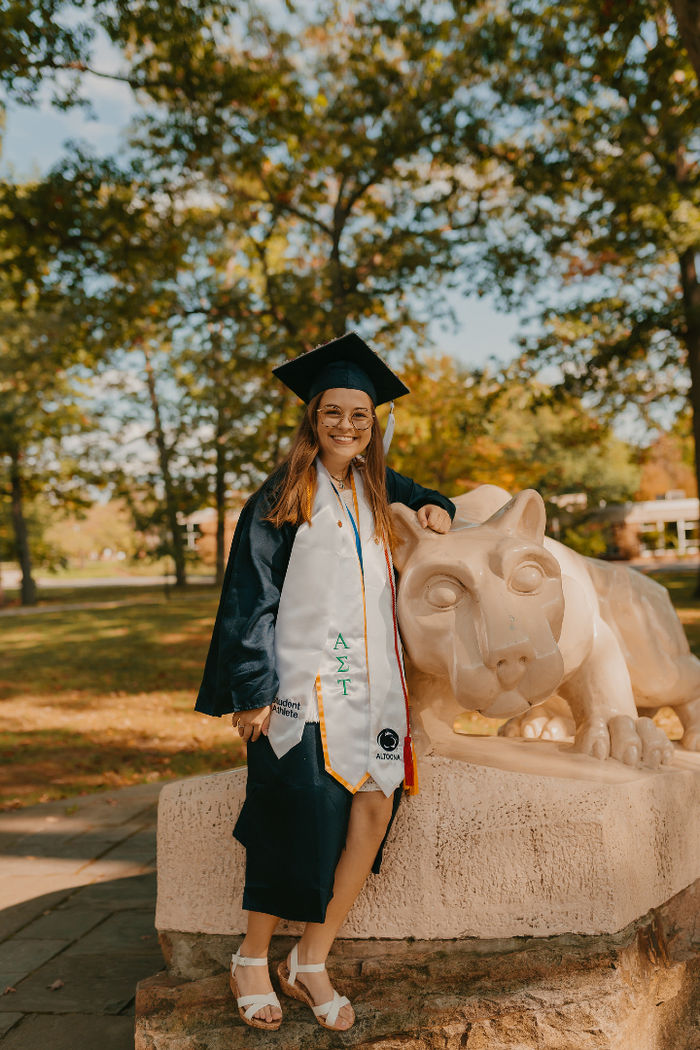 Woman poses in graduation cap and gown with a Lion Shrine statue