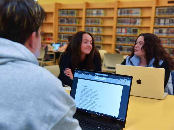 Three students sit at a table in a library, engaged in conversation. Two students face the camera, one with a MacBook, while the third has their back to the camera with a laptop displaying a Word document. Bookshelves filled with books line the background, indicating a quiet academic environment.