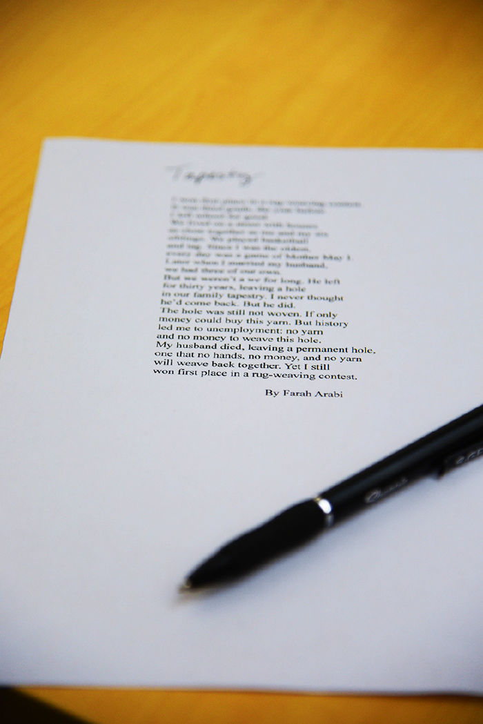 A close-up of a printed poem titled Tapestry by Farah Arabi sits on a wooden table. A black pen rests diagonally across the lower corner of the paper. The poem discusses themes of loss, resilience, and weaving as metaphor, with part of the text clearly legible.