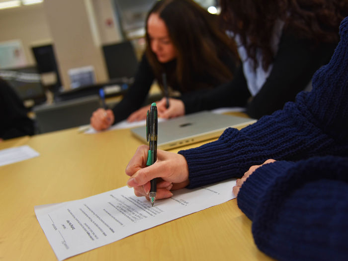 Close-up of students writing on printed worksheets at a table. One hand in the foreground holds a pen, poised to write. In the background, two students focus on their work, with a laptop partially visible between them. The setting appears to be a classroom or library study area.