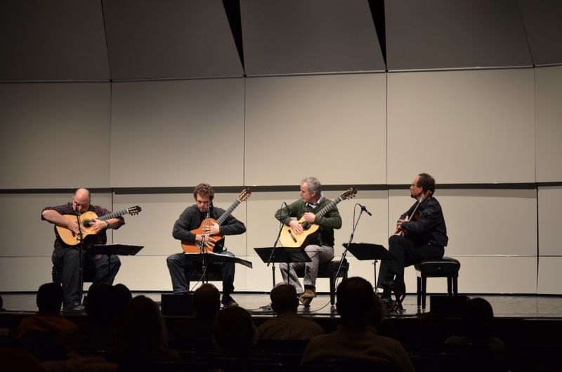 Four guitarists sit in chairs and perform on stage.