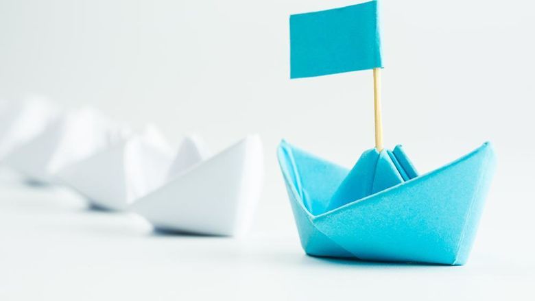 a blue paper sailboat leading other white paper sailboats