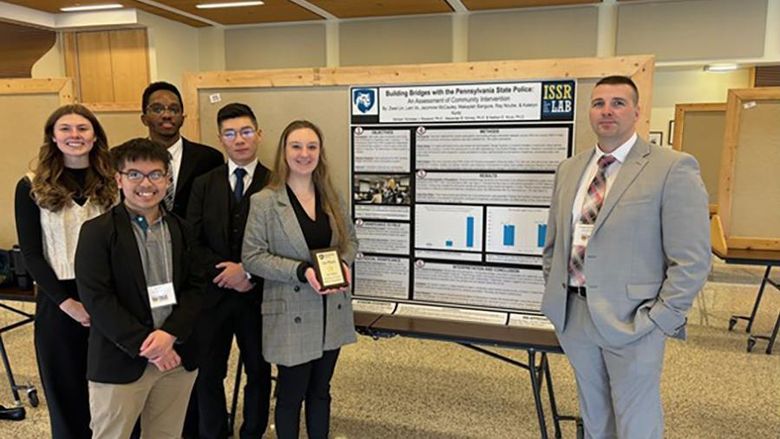 Penn State Altoona students Jazzmine McCauley, Ziwei (Will) Lin, Ray Ncube, Lam Vo, and Katelyn Kurtz standing next to their first-place poster, “Building Bridges with Pennsylvania State Police: An Assessment of Community Outreach,” and Trooper Joe Dunsmore, keynote speaker of the 2024 PACJE conference.  