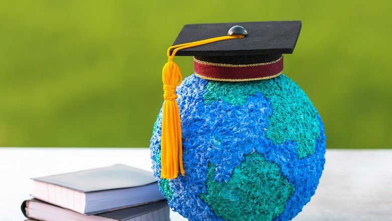 A small globe with a graduation cap on it sitting next to a pile of books