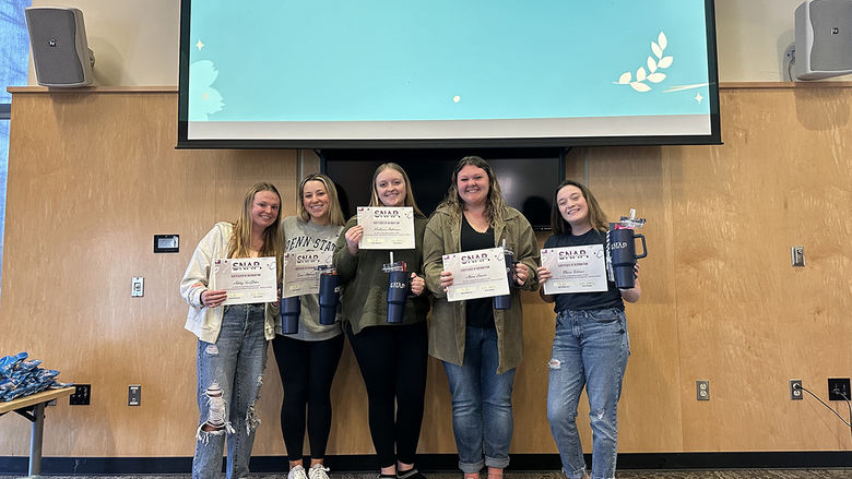 A group of five young women stand together in front of a presentation screen that reads Committee Chairs! Each of them holds a certificate and a tumbler, smiling proudly. The setting appears to be a conference or meeting room, with a professional yet relaxed atmosphere. They are dressed casually, with some wearing Penn State apparel, indicating a possible university affiliation.