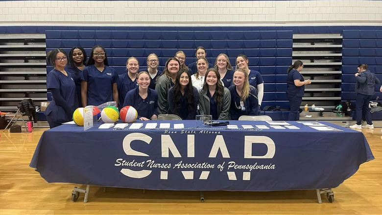 A group of student nurses from the Student Nurses Association of Pennsylvania (SNAP) at Penn State Altoona pose together behind an informational table in a gymnasium. They are dressed in navy blue scrubs and are smiling at the camera. The table is covered with a SNAP-branded tablecloth and displays brochures, beach balls, and other materials. In the background, additional students and attendees can be seen engaging in activities, suggesting an event or fair setting.