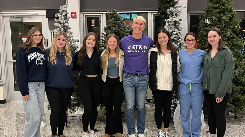 A group of eight students pose together in a well-lit indoor space decorated with Christmas trees, some with snow-like flocking. They are dressed casually, with one student wearing blue medical scrubs and another wearing a SNAP (Student Nurses Association of Pennsylvania) t-shirt. The students are smiling, standing close together, and appear to be celebrating or attending a seasonal event.