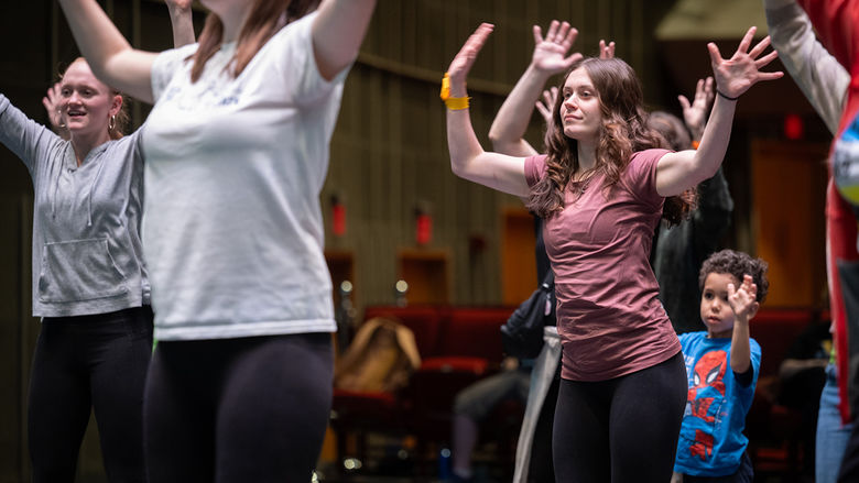 A group of young people dance on a stage. One girl is in focus with her hands waving in the air