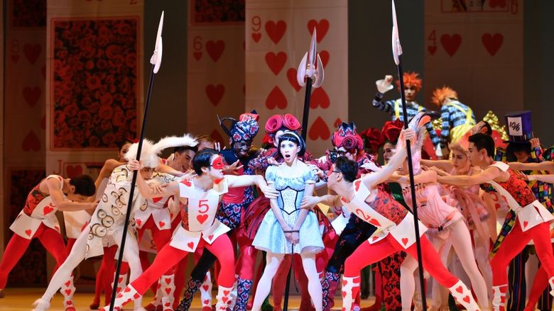 A dancer resembling Alice is surrounded by dancers dressed as playing cards and flamingoes.