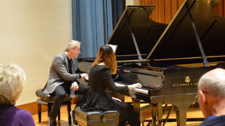 A man and woman each sit at their own piano while he coaches the student musician.