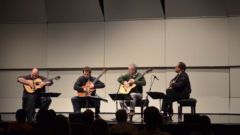 Four guitarists sit in chairs and perform on stage.
