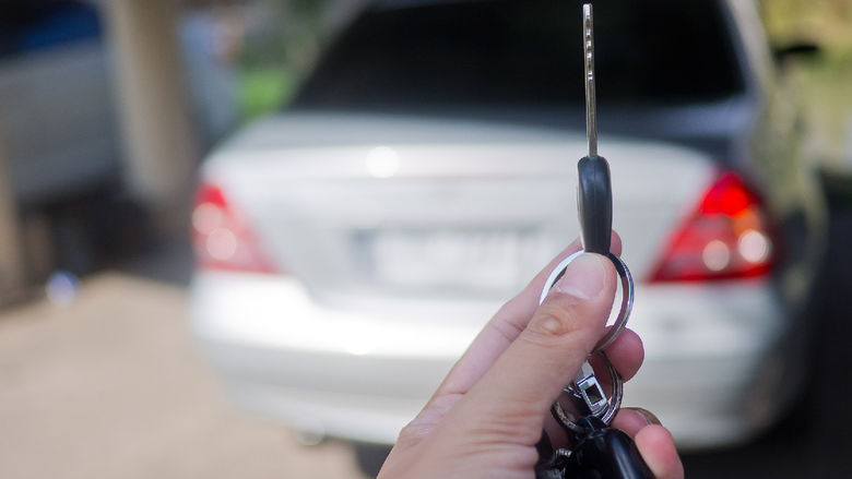 A hand holding up a car key in the foreground with a motor vehicle in the background