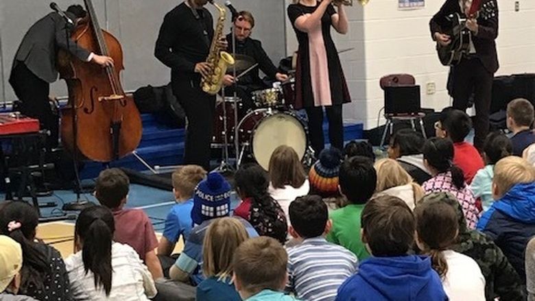 Musicians stand while they perform in front of a group of middle-school-aged students sitting on a floor.