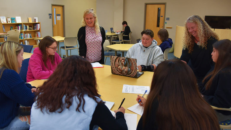 A group of students sits around a table in a library or classroom setting, engaged in discussion and writing. Two instructors stand with them, smiling and interacting. Bookshelves, chairs, and a clock are visible in the background, suggesting an academic or collaborative learning environment.