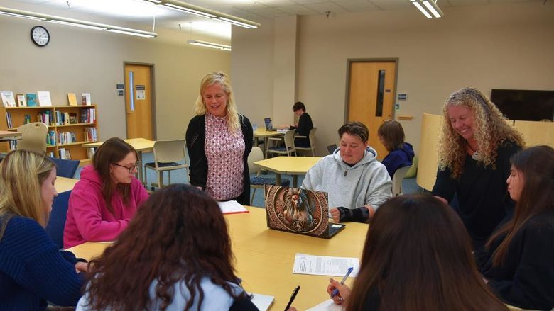 A group of students sits around a table in a library or classroom setting, engaged in discussion and writing. Two instructors stand with them, smiling and interacting. Bookshelves, chairs, and a clock are visible in the background, suggesting an academic or collaborative learning environment.