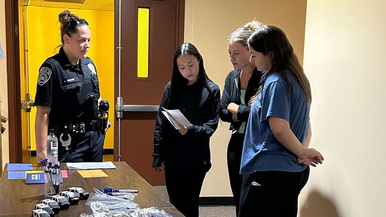 A police officer speaks with three women who are reading from a pamphlet at a table with promotional items in a hallway.
