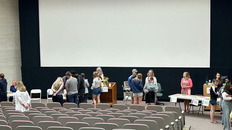 People gather at various tables in a large lecture hall, engaging with exhibitors beneath a large blank screen.