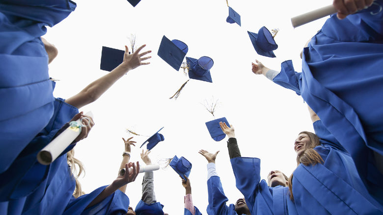 A group of students wearing blue graduation robes throw their caps in the air