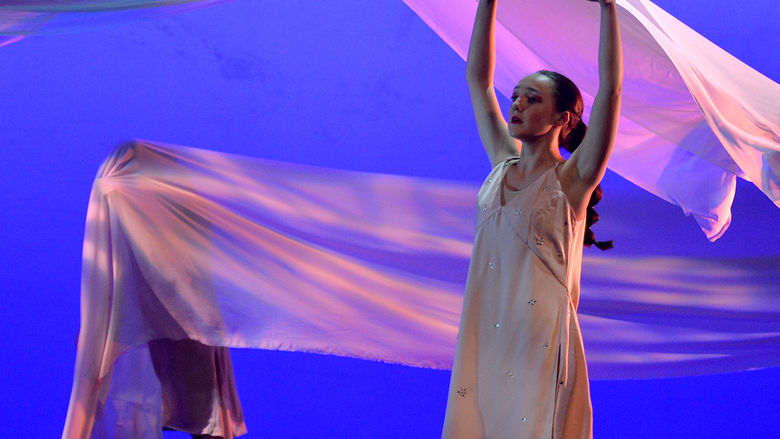 A dancer in the foreground with her arms raised in the air while a piece of fabric flows in the air behind her