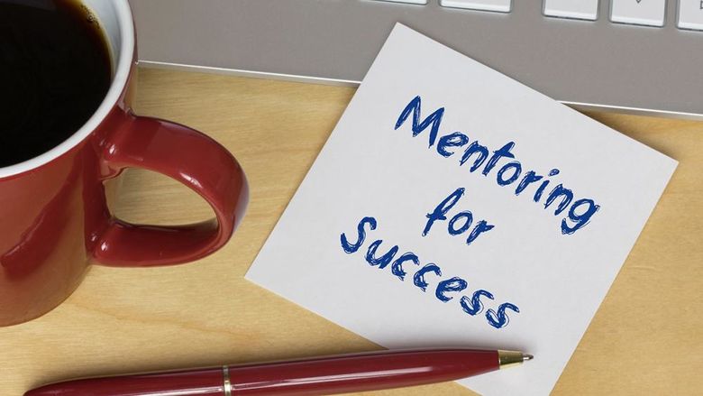 A post-it note reading Mentoring for Success sits on a desk with a pan, a cup of coffee, and a laptop