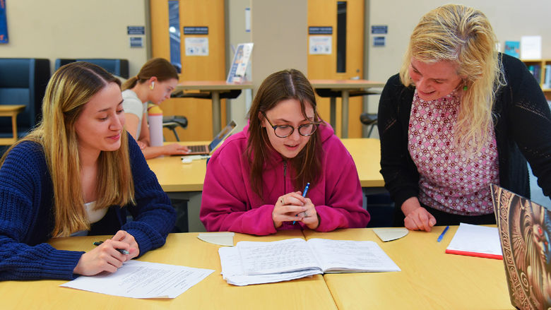 A student in a pink hoodie reads from an open workbook while seated between another student and an instructor. The instructor leans in to offer support, smiling. A third student listens attentively, holding a pen. The setting appears to be a library or study space with bookshelves and computers in the background.