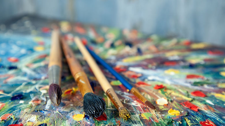 This close-up photo shows paintbrushes resting on a vibrant, abstract palette with thick dabs of colorful paint, evoking a lively artistic atmosphere.