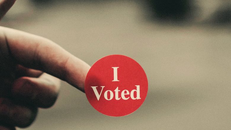 closeup image of person's hand holding red "I Voted" sticker on fingertip