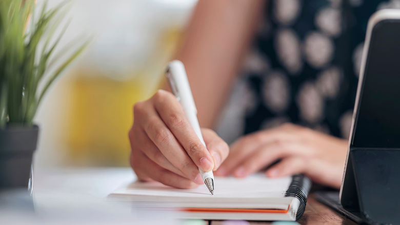 The hands of a woman writing on a notepad with a laptop computer sitting nearby