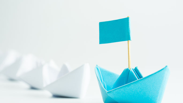 a blue paper sailboat leading other white paper sailboats