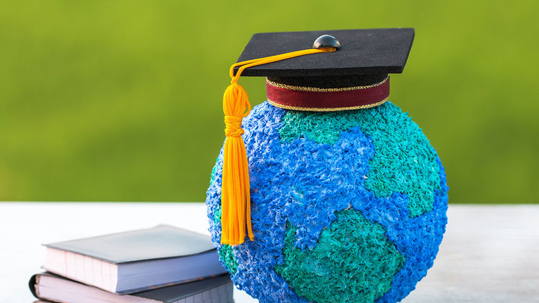 A small globe with a graduation cap on it sitting next to a pile of books