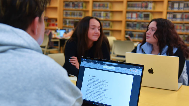 Three students sit at a table in a library, engaged in conversation. Two students face the camera, one with a MacBook, while the third has their back to the camera with a laptop displaying a Word document. Bookshelves filled with books line the background, indicating a quiet academic environment.