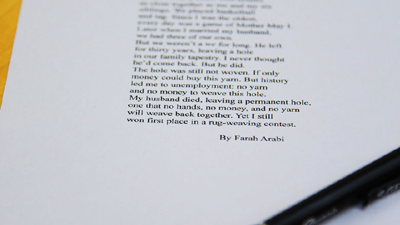 A close-up of a printed poem titled Tapestry by Farah Arabi sits on a wooden table. A black pen rests diagonally across the lower corner of the paper. The poem discusses themes of loss, resilience, and weaving as metaphor, with part of the text clearly legible.
