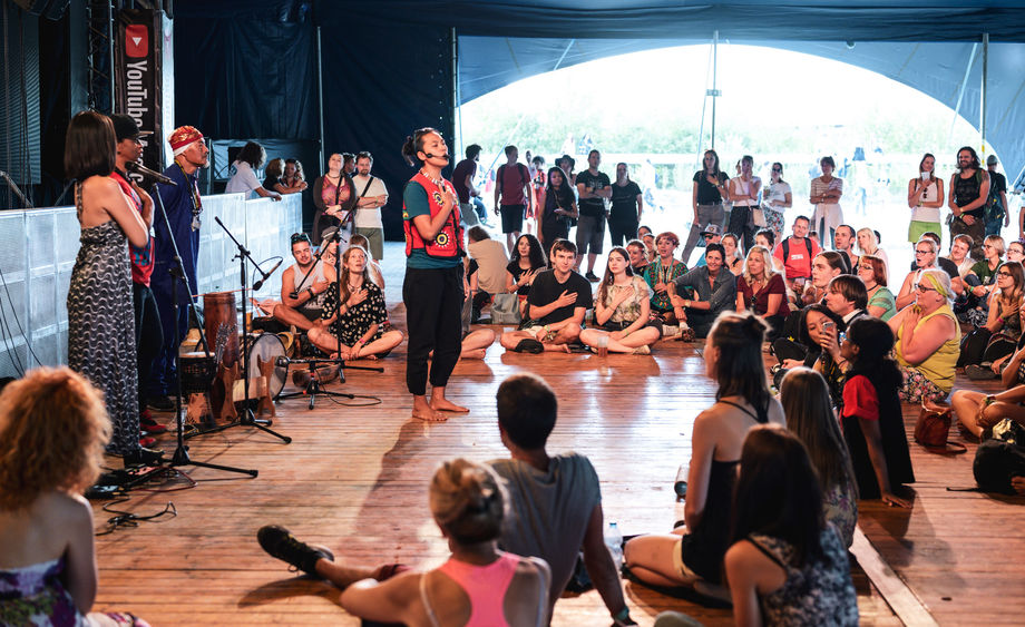 Four people stand in front of microphones and sing in front of a diverse crowd seated in front of them.