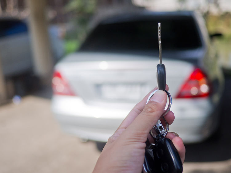 A hand holding up a car key in the foreground with a motor vehicle in the background